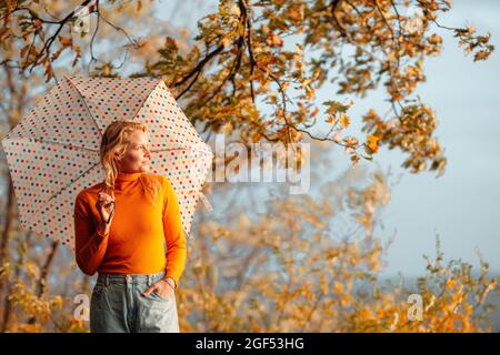 Donna che guarda via mentre si alza in piedi con la mano in tasca che tiene l'ombrello il giorno di sole Foto Stock