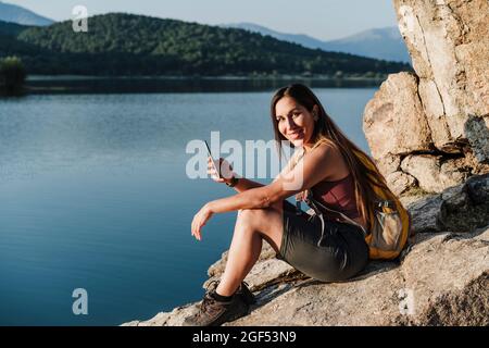 Sorridente escursionista donna che tiene il telefono cellulare mentre si siede sulla roccia vicino al lago Foto Stock