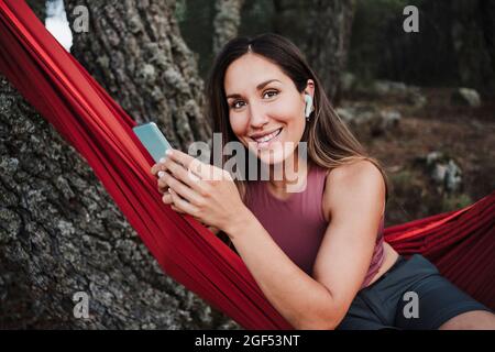 Giovane donna sorridente con telefono cellulare che ascolta musica mentre si siede su amaca Foto Stock