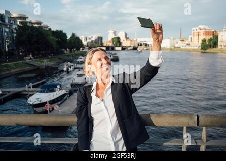 Donna d'affari sorridente che prende selfie attraverso lo smartphone al fiume Foto Stock