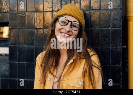 Donna rossa sorridente che indossa una giacca gialla mentre si appoggia sulla parete nera Foto Stock