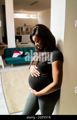 Donna incinta sorridente che tocca l'addome mentre si appoggia contro la colonna Foto Stock