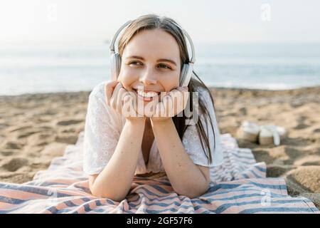 Donna sorridente con mani sul mento che ascolta musica in spiaggia Foto Stock