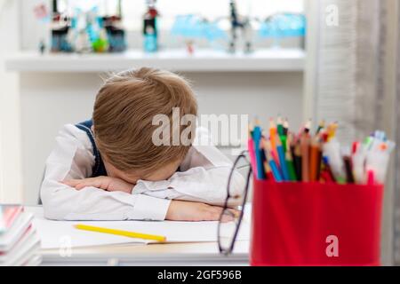 Un ragazzo carino primo grader in una divisa della scuola a casa durante una pandemia si addormentò facendo i compiti ad una scrivania con libri e matite. Messa a fuoco selettiva. Cl Foto Stock