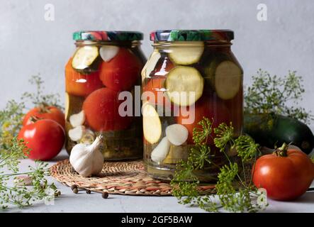 Zucchine marinate con pomodori in due vasetti di vetro per l'inverno su sfondo chiaro con ingredienti freschi Foto Stock