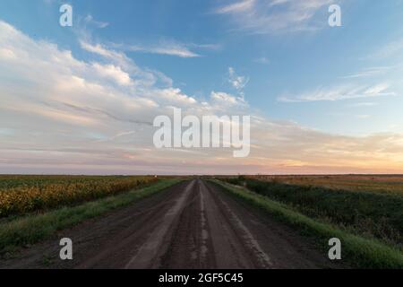 Un ampio angolo di paesaggio girato di una strada sterrata aperta tra campi agricoli con un cielo drammatico e le nuvole arancione e blu al tramonto Foto Stock