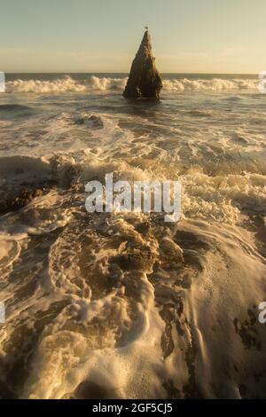 Un ampio angolo verticale di onde bianche che si infrangono sulla spiaggia sabbiosa di una spiaggia con un alto punto di roccia al largo della costa con un uccello arroccato sulla cima Foto Stock