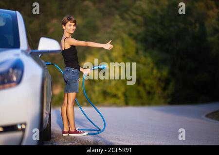 Ragazza in piedi accanto all'auto elettrica tenendo un cavo di ricarica e hitchhiking. Foto Stock