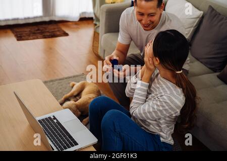 Giovane uomo che propone la sua ragazza con anello di fidanzamento a casa. Donna asiatica che si sorprende dal suo ragazzo con la proposta di matrimonio mentre lavora su un laptop Foto Stock