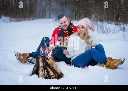 una giovane coppia, un uomo adulto e una donna, si siedono nella neve vicino ad un fuoco fuori in inverno. I giovani indossano jeans, maglioni e scarpe invernali. Inverno nuvoloso giorno, alberi sullo sfondo. Foto Stock