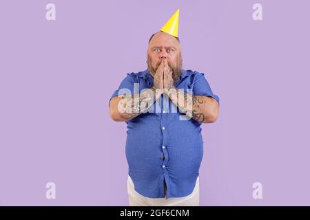 Uomo sorpreso con sovrappeso in camicia stretta tiene le mani sulla bocca su sfondo viola Foto Stock