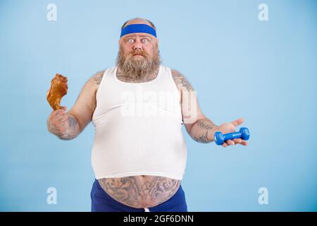 L'uomo confuso con sovrappeso sceglie tra coscia di pollo e manubra su sfondo blu chiaro Foto Stock