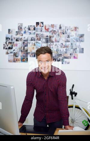 Ritratto di un bel giovane e sorridente designer grafico in camicia a quadri appoggiati alla scrivania con computer e tablet grafico e sorridenti alla fotocamera Foto Stock
