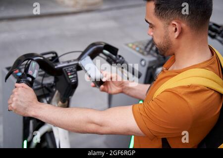 Il giovane uomo latino si alza accanto al parcheggio con una bicicletta a noleggio e usa uno smartphone con un sorriso sul viso. Concetto di trasporto ecologico. Foto di alta qualità Foto Stock