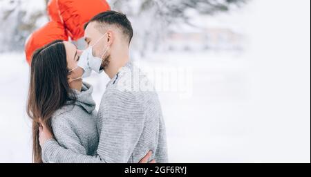 Giovane coppia innamorata nel parco su uno sfondo di neve e palloncini rossi a forma di cuore. Regalo di San Valentino. Gli amanti baciano in maschere mediche Foto Stock