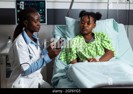 Donna con professione medica che dà la medicina di prescrizione a paziente malato che posa in letto di reparto dell'ospedale. Bottiglia di medico afro-americano contenente pillole, farmaci, trattamento per la malattia Foto Stock