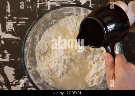 Donna mano tenendo la siviera con acqua calda e versandola nella farina per fare l'impasto sul fondo marrone Foto Stock