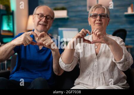 Famiglia anziana romantica che crea il simbolo della forma del cuore utilizzando le mani per la fotocamera a casa. Vecchio marito sposato e moglie seduti insieme sul divano facendo amore concetto gesto gesto sentimento allegro Foto Stock