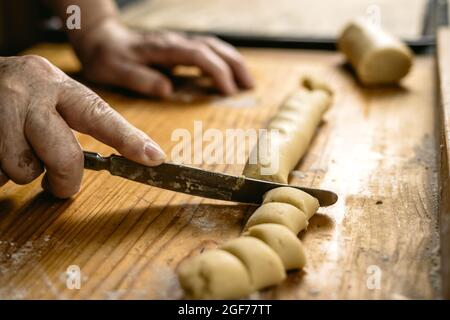 Le vecchie mani tagliano la pasta di pasticceria con il coltello su un tavolo di legno. Donna anziana che fa biscotti fatti in casa. Preparazione del cibo Foto Stock