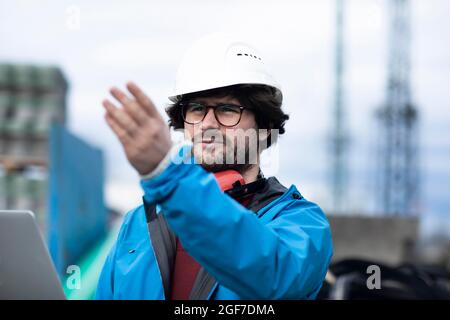 Giovane ingegnere che indossa un casco e una protezione acustica sul posto di lavoro all'esterno con un computer portatile, Friburgo, Baden-Wuerttemberg, Germania Foto Stock