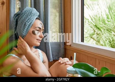 Routine di cura della pelle. Donna di mezza età con rughe e idrogel sotto gli occhi cerotti bere frullato e guardare in finestra. Sollevamento maschera antirughe und Foto Stock