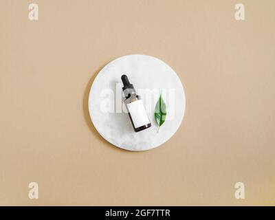 Podio rotondo bianco marmo, pipetta in bottiglia color ambra con etichetta bianca e foglia verde su sfondo beige. Vista dall'alto. Spazio di copia Foto Stock