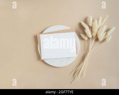 Carta bianca bianca per testo e busta sul podio rotondo in marmo accanto al bouquet di Lagurus secco su sfondo beige. Concetto minimo per messaggio, messaggio Foto Stock