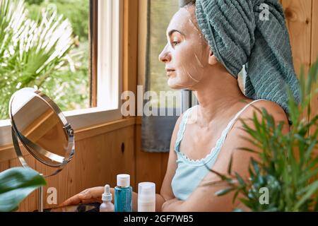 Routine di cura della pelle. Donna di mezza età con foglio maschera idratante nel suo viso guarda attraverso la finestra. Sollevamento della maschera antirughe sotto gli occhi. Collagene m Foto Stock