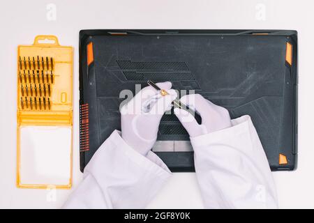 Vista dall'alto di un tecnico con un guanto bianco, sollevando un cacciavite per aprire il coperchio di un computer portatile prima di essere riparato su un tavolo bianco Foto Stock