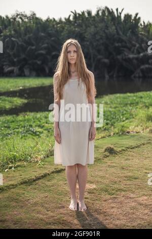 Ritratto a tutta lunghezza di giovane graziosa donna caucasica in abito bianco in piedi a piedi nudi nel parco. Capelli biondi lunghi. Bellezza naturale. Il make-up è gratuito. Foto Stock