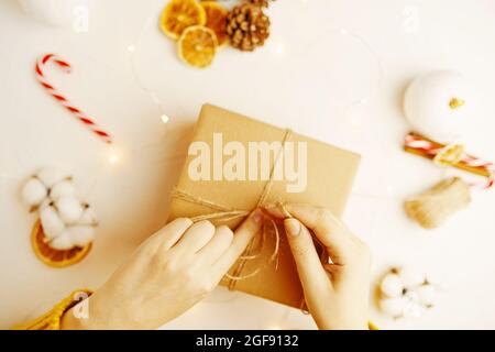 Confezione della confezione regalo di Natale. Coni di abete, fette di arancio secco e altre decorazioni sul tavolo. Ragazza in maglione sta preparando sorpresa per l'anno nuovo. Atmosfera festosa. Nastro di legatura per le mani delle donne in nodo. Foto Stock