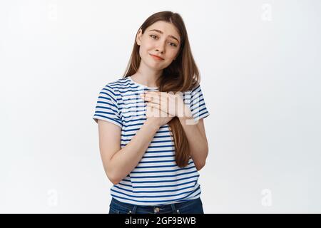 Immagine di una giovane donna toccata che tiene le mani sul cuore, sospira e guarda con affetto e amore, sfondo bianco Foto Stock