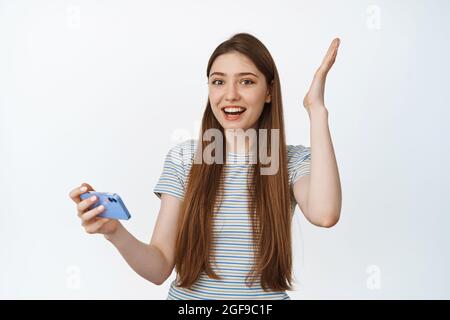 La ragazza felice balla, tenendo lo smartphone e sorridendo stupito, vincendo sul cellulare, in piedi su sfondo bianco Foto Stock