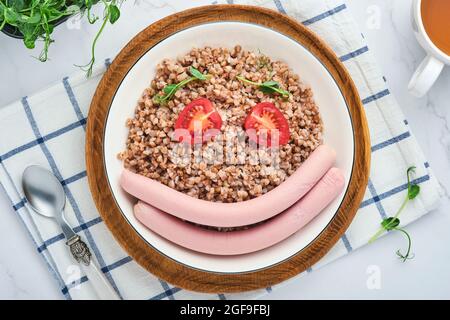 Porridge di grano saraceno bollito, salsicce di carne, pomodoro e micrograni di piselli in un piatto bianco su fondo grigio pietra. Divertente idea alimentare arte pranzo sano fo Foto Stock