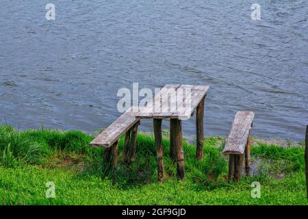 Tavolo e panchine in legno sulla costa del lago. Erba verde e calmo superficie d'acqua. Area relax. Foto Stock