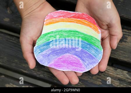 Le mani di un bambino piccolo stanno tenendo delicatamente un pezzo di carta arcobaleno disegnato con matite colorate. Foto Stock