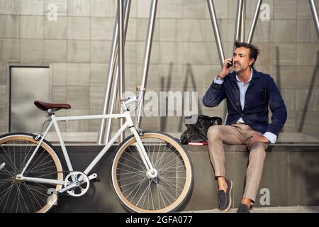 Uomo adulto seduto a parlare al telefono con la bicicletta appoggiata al muro. Concetto di trasporto ecologico. Foto di alta qualità Foto Stock