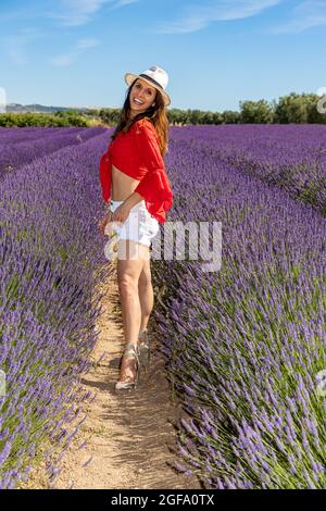 Una giovane donna sorridente che si posa in un campo di lavanda in fiore. Indossa una blusa rossa, pantaloncini e un cappello bianco. Concetto di benessere e amore per nat Foto Stock