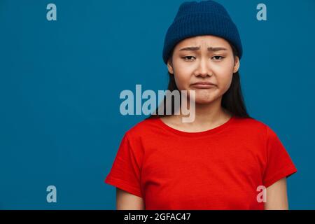 Sconvolto giovane donna asiatica triste in abiti casual in piedi su sfondo blu parete, piangendo Foto Stock