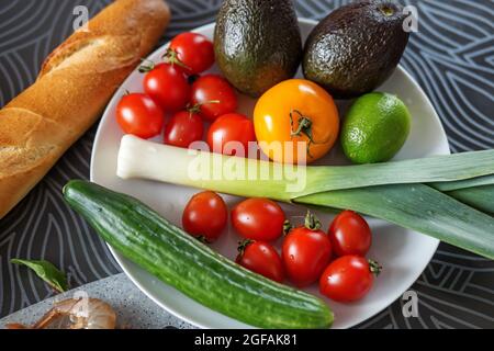 Verdure fresche su un piatto bianco, avocado, scalogni, pomodori, cetrioli. Per il giorno del vegetarianismo Foto Stock