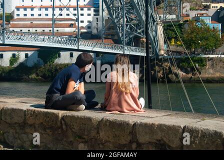 Una coppia è seduta all'imbarco del fiume Douro parlando, un ponte sullo sfondo - Portogallo, Porto Foto Stock