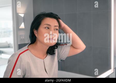 Perdita di capelli matura donna asiatica che tocca i suoi capelli styling o colorazione capelli grigi guardando se stessa nello specchio di casa. Beautifiul signora professionale cinese Foto Stock