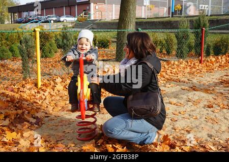 POZNAN, POLONIA - 01 novembre 2015: Una giovane mummia inginocchiata da un ragazzo seduto su un'altalena primaverile in un parco giochi in autunno Foto Stock