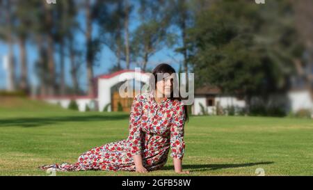 Ritratto di bella giovane donna latina guardando a destra con abito floreale di design poggiato sull'erba in mezzo ad un giardino con sfondo Foto Stock