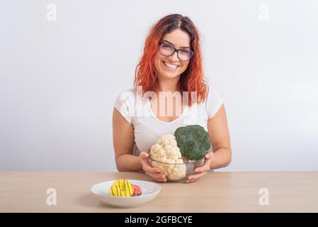 La donna caucasica preferisce cibo sano. Ragazza rossa sceglie tra broccoli e ciambelle su sfondo bianco. Foto Stock