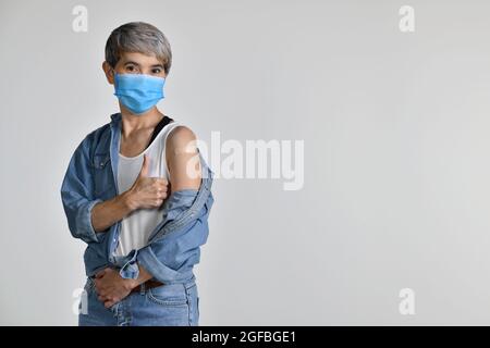 Donna asiatica di mezza età felice vaccinata mostrando i pollici in su al cerotto adesivo sul suo braccio dopo l'iniezione antivirale di coronavirus isolato su wh Foto Stock