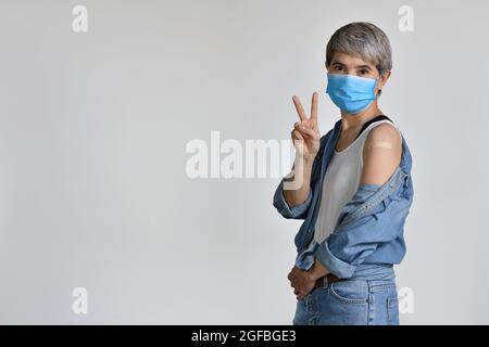 Donna asiatica di mezza età felice che indossa maschera chirurgica per il virus corona e vaccinato sorridendo con il volto felice alla macchina fotografica facendo segno di vittoria mostrando un Foto Stock
