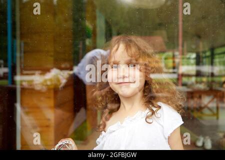 Ragazza felice carina con capelli ricci sorridenti e guardando la macchina fotografica mentre si trova dietro vetro trasparente Foto Stock