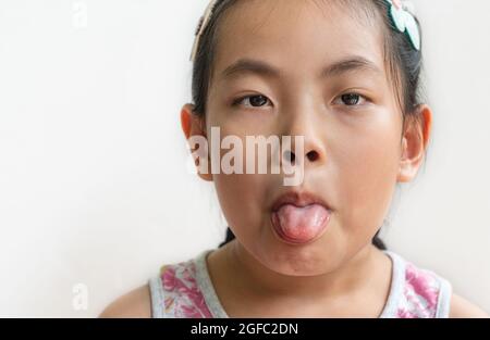 Headshot di ragazza carina asiatica con un volto grimming, capelli lunghi neri legati, guarda e mostra lingua fuori alla macchina fotografica, si erge su parete bianca Foto Stock