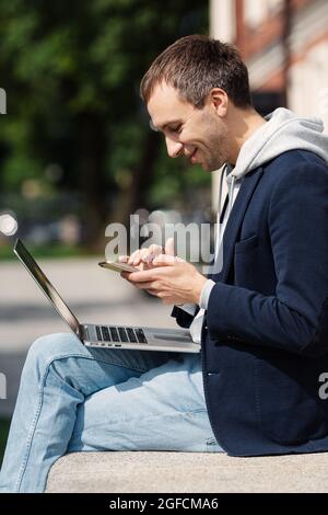 Un uomo d'affari sorridente e felice ha letto la notifica delle app nello smartphone che lavora su un notebook in remoto all'aperto Foto Stock